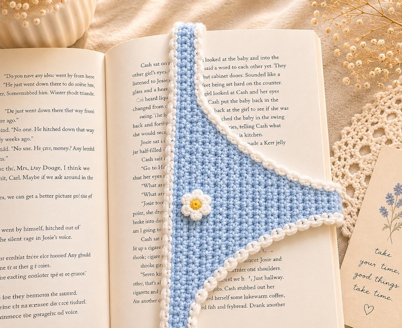 May include: A handmade, light blue crochet bookmark with a white scalloped edge and a small white and yellow daisy. The bookmark is placed in an open book, adding a touch of handmade charm to reading.
