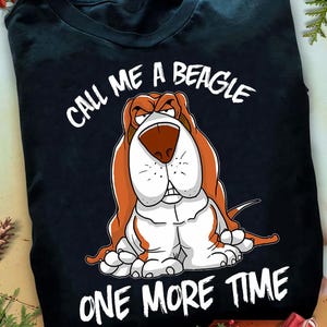 以下が含まれることがあります： 不機嫌そうな表情のビーグル犬の漫画が描かれた黒いTシャツ。ビーグル犬は茶色、白、タン色です。「CALL ME A BEAGLE ONE MORE TIME」というテキストが画像の上部と下部に白で印刷されています。