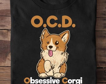 Op de afbeelding: Zwart T-shirt met een cartoon corgi. De corgi heeft bruin en wit haar en een roze tong. De tekst "O.C.D. Obsessive Corgi Disorder" staat in oranje en wit. Het shirt is op een houten oppervlak gevouwen.