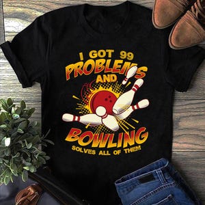 May include: Black t-shirt with a bowling-themed graphic. The design features a red bowling ball, white bowling pins, and the text "I GOT 99 PROBLEMS AND BOWLING SOLVES ALL OF THEM" in yellow and red.