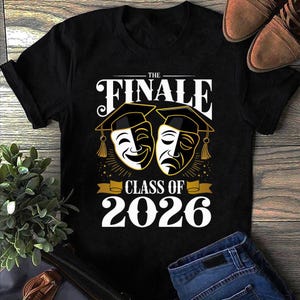 May include: Black t-shirt with the text "THE FINALE CLASS OF 2026". The design features two theatre masks, one smiling and one frowning, each wearing a graduation cap. The shirt is a crew neck and has short sleeves.