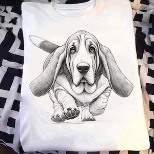 May include: White t-shirt with a detailed black and white illustration of a Basset Hound dog. The dog is depicted in motion, with long ears and a wrinkled face. The illustration gives the impression of movement. The shirt is made of a soft material.