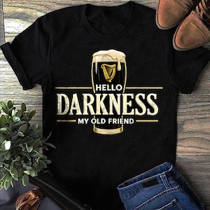 May include: A black short-sleeve t-shirt featuring a graphic of a pint glass filled with dark liquid and foam. The text reads "HELLO DARKNESS MY OLD FRIEND" in a distressed white font, with a gold harp icon centered above the word "HELLO".