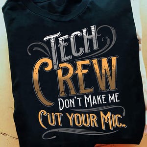 May include: Black t-shirt with the words "TECH CREW" in white and orange lettering. The text "DON'T MAKE ME CUT YOUR MIC" is below the main text. The shirt is folded on a light-colored surface.