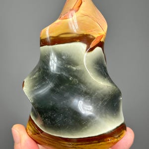 May include: A polished, abstract stone sculpture with a layered appearance. The colors include shades of tan, brown, white, and dark green. The top is a pointed shape, and the base is rounded. The stone has a glossy finish.