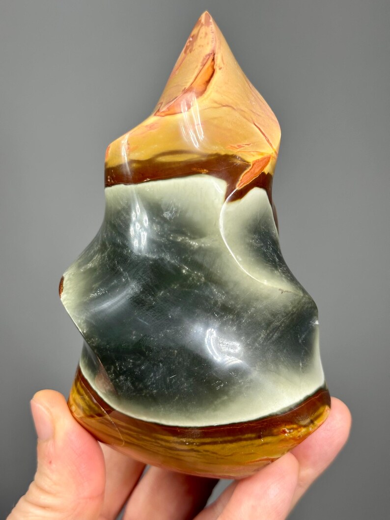 May include: A polished, abstract stone sculpture with a layered appearance. The colors include shades of tan, brown, white, and dark green. The top is a pointed shape, and the base is rounded. The stone has a glossy finish.