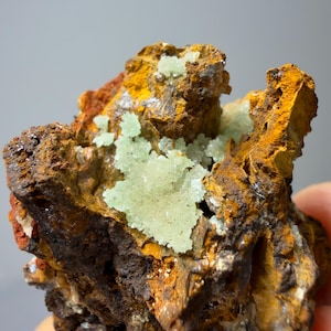 May include: A close-up of a mineral specimen featuring a cluster of light green crystals on a matrix of brown, orange, and yellow rock. The rock has a rough, textured surface with areas of dark brown and reddish-brown.