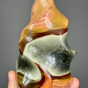 May include: A polished, abstract stone sculpture with a flame-like shape. The stone features bands of color, including shades of orange, brown, white, and black. The sculpture is held in a hand, showcasing its size and texture.