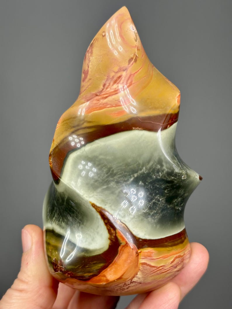 May include: A polished, abstract stone sculpture with a flame-like shape. The stone features bands of color, including shades of orange, brown, white, and black. The sculpture is held in a hand, showcasing its size and texture.
