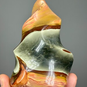 May include: A polished, abstract stone sculpture with a flame-like shape. The stone features bands of color, including orange, brown, white, and gray. The sculpture has a smooth, glossy finish and is held in a hand against a gray background.