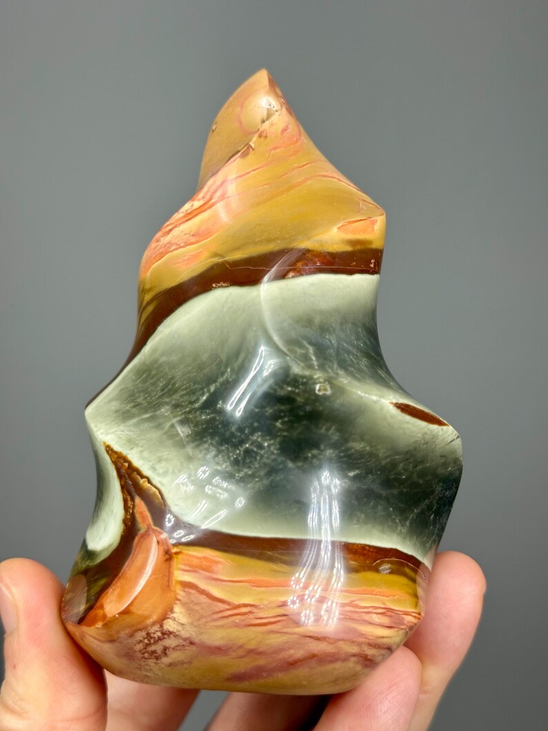 May include: A polished, abstract stone sculpture with a flame-like shape. The stone features bands of color, including orange, brown, white, and gray. The sculpture has a smooth, glossy finish and is held in a hand against a gray background.