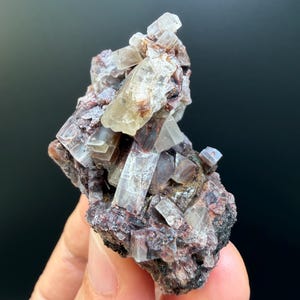 May include: A raw mineral specimen featuring translucent, pale, prismatic crystals clustered atop a dark, textured, reddish-brown matrix. The crystalline structure displays sharp, geometric edges and a glassy luster, contrasting with the rough, earthy base of the stone formation.