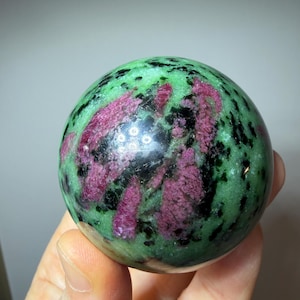 Peut inclure: Une sphère de pierre précieuse polie, présentant un mélange de couleurs vives. La sphère est principalement verte, avec des inclusions magenta et noires. La surface lisse reflète la lumière, mettant en valeur les motifs et textures naturels de la pierre.