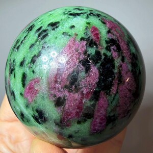 May include: A polished spherical stone featuring a vibrant mix of bright green zoisite, deep magenta ruby inclusions, and scattered black hornblende crystals. The surface is smooth and reflective, showcasing a natural, multi-colored mineral pattern.