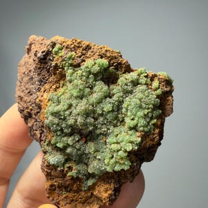 May include: A rough, brown rock formation with a cluster of small, light green crystals. The crystals have a textured, granular appearance. The rock is held in a hand against a neutral background.