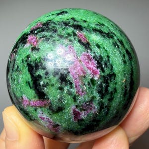 Peut inclure: Une pierre sphérique polie présentant une base de couleur vert vif avec des inclusions minérales noires proéminentes et des taches irrégulières dispersées de rose magenta profond sur toute la surface, créant un aspect multicolore contrasté.