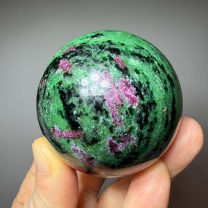May include: A polished sphere of green zoisite with ruby inclusions. The gemstone features a vibrant green base color with black and pinkish-red streaks and spots. The sphere is held in a hand against a neutral background.