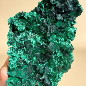 May include: A raw, natural malachite specimen. The mineral features a deep green color with a textured, crystalline surface. The stone has areas of dark green and black, with a rough, uneven shape. The malachite is a natural gemstone.