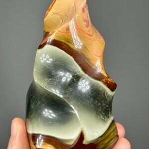 May include: A polished, abstract stone sculpture with a flowing, organic shape. The stone features bands of color, including tan, brown, green, and white. The sculpture is held in a hand, showcasing its size and texture.