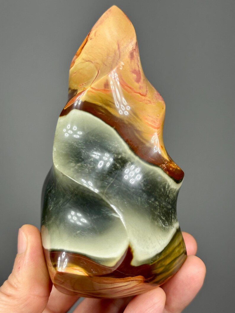 May include: A polished, abstract stone sculpture with a flowing, organic shape. The stone features bands of color, including tan, brown, green, and white. The sculpture is held in a hand, showcasing its size and texture.