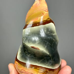 May include: A polished, abstract sculpture in shades of orange, brown, black, and cream. The stone has a swirling, layered pattern and a smooth, glossy finish. The sculpture is held in a hand, showcasing its unique shape and color variations.