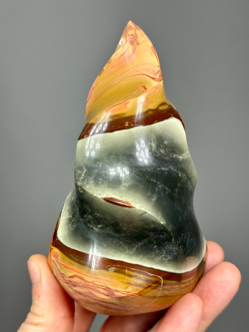 May include: A polished, abstract sculpture in shades of orange, brown, black, and cream. The stone has a swirling, layered pattern and a smooth, glossy finish. The sculpture is held in a hand, showcasing its unique shape and color variations.