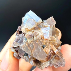 May include: A raw mineral specimen featuring a cluster of translucent, light brown and tan crystals with a glossy, layered texture. The formation includes a prominent, pale blue-tinted crystal at the top, with small dark inclusions embedded throughout.