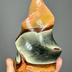 May include: A polished, freeform stone sculpture with a layered appearance. The stone features bands of tan, orange, brown, and gray hues. The sculpture has a smooth, glossy finish and is held in a hand.