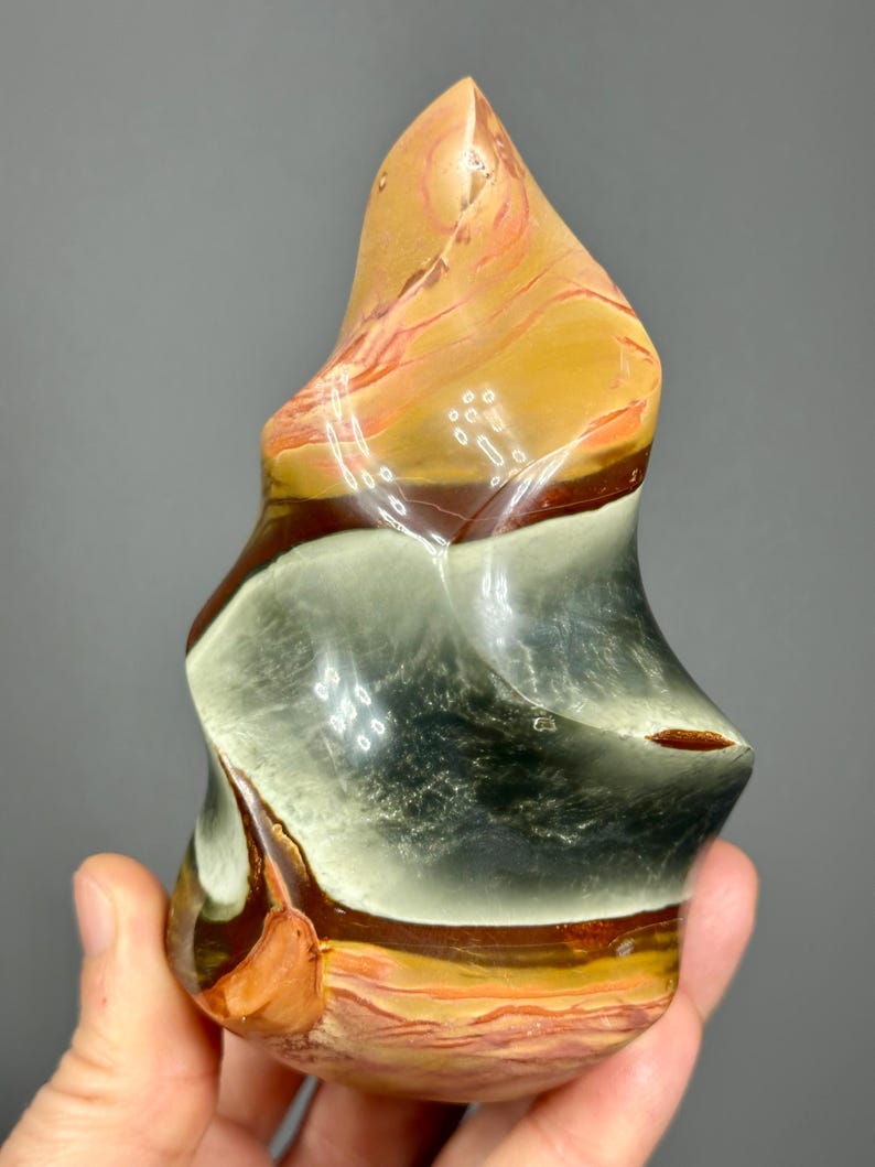 May include: A polished, freeform stone sculpture with a layered appearance. The stone features bands of tan, orange, brown, and gray hues. The sculpture has a smooth, glossy finish and is held in a hand.