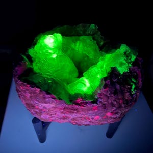 May include: A glowing green mineral formation inside a pink and purple rock structure. The mineral has a bright, almost neon, appearance. The rock structure is supported by three dark legs, and the background is black.