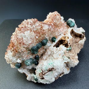 Peut inclure: Gros plan sur un spécimen minéral présentant un amas de cristaux de quartz transparents avec des inclusions brun rougeâtre. Des formations de malachite sphériques vert foncé sont dispersées sur la surface. Le minéral est posé sur un fond sombre.