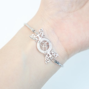 Pode incluir: Uma pulseira de prata com um design de nó celta. A pulseira tem uma peça central circular com um padrão de nó detalhado, rodeada por pequenas pedras brilhantes. A pulseira é usada em um pulso, mostrando seu design intrincado e estilo elegante.