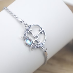 May include: A silver-toned bracelet featuring a circular pendant with an intricate openwork design. The pendant includes a central cross-like symbol, crescent moon accents, and a small, round, iridescent cabochon stone set within the metal frame.