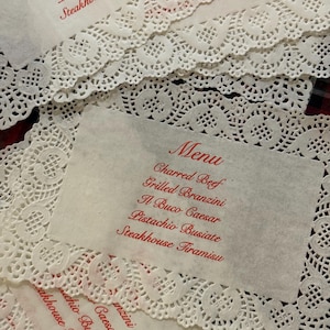 May include: Several white paper menus with red text and decorative lace edges are arranged on a surface. The menus list various food items, including "Charred Beef" and "Steakhouse Tiramisu."