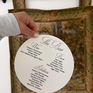 May include: A white, circular bar menu with the text "The Bar" at the top. The menu lists wine, beer, and libations, including bourbon, tequila, and gin. The menu is held in front of a decorative tapestry.