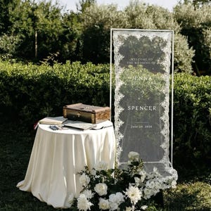 May include: Wedding welcome sign with white lace trim, featuring the names Jenna and Spencer, and the date June 20, 2026. A small table with a white tablecloth holds a wooden box and guest book. White flowers decorate the base of the sign.