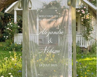 May include: A rectangular white sheer fabric wedding sign with lace trim, hanging from a tree branch by two sage green satin ribbons. The text reads: WELCOME TO THE WEDDING OF Alejandro & Nohemi 18 JULY 2025, printed in white lettering.