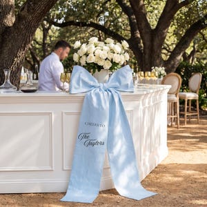 May include: A white bar decorated with a large light blue bow and a floral arrangement of white roses. The bow has the text "Cheers to The Taylors" printed on it. The bar is set up outdoors with a bartender in the background.