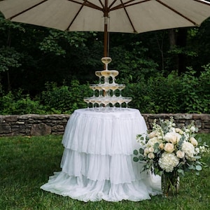 May include: A tiered champagne coupe tower sits on a round table draped in a white ruffled fabric tablecloth. A large floral arrangement with white roses and greenery stands beside the table under a large beige patio umbrella outdoors.
