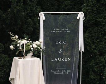 May include: A wedding welcome sign with the text "Welcome to the Wedding of Eric & Lauren" and the date September 6th, 2026, Plymouth, NH. The sign is white and draped with a sheer fabric. A table with a floral arrangement and candles is to the left.