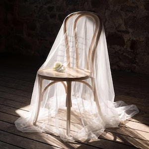 May include: A light-coloured wooden bentwood chair draped in a sheer white veil featuring printed cursive text. A single white flower rests on the circular seat of the chair. The fabric cascades down from the backrest to the floor in soft folds.