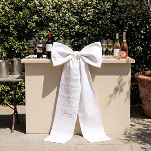 May include: An outdoor bar in cream with a large white bow featuring drink options. The bar is set with wine bottles, glasses, and a champagne bottle. The bow has text that reads "Site Cuple", "Wine & Bubbles", "No Alcohol", and "Cheers!"