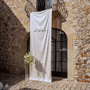 May include: A long, off-white fabric banner hanging vertically from a balcony, featuring the text "the Malones" in a dark, elegant script font. A clear acrylic pedestal stands to the left, topped with a large arrangement of green and white flowers.