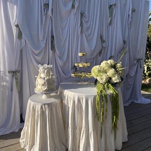 May include: A white tiered cake on a pedestal stand, a multi-level glass dessert tower, and a large bouquet of white hydrangeas with green hanging amaranthus on a round table, all set against a white draped fabric backdrop with sage green ribbons.