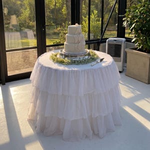 May include: A three-tiered white cake sits on a silver pedestal atop a round table draped in a white, multi-layered ruffled fabric tablecloth. A ring of green foliage encircles the base of the cake, and a silver cake server rests on the table.