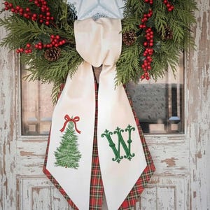 Peut inclure: Une couronne de fête composée de branches de cèdre vertes, de baies rouges et de pommes de pin brunes. Un ruban en tissu crème pend du centre, affichant un sapin de Noël vert brodé avec un nœud rouge et une grande lettre W verte.