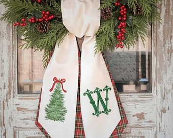 May include: A decorative fabric hanging featuring a cream-colored front with an embroidered green Christmas tree topped with a red bow and a green embroidered letter W. The back layer is a red, green, and gold plaid fabric with pointed ends.