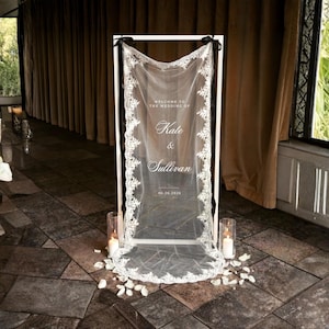 Può includere: Un cartello nuziale con cornice bianca con un pannello in tessuto trasparente. Il pannello è adornato con finiture in pizzo bianco e presenta il testo "Welcome to the Wedding of Kate & Sullivan 06.26.2026". Due candele in portacandele di vetro fiancheggiano il cartello.