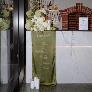 Może przedstawiać: Bar z marmurowym blatem i zielonym satynowym menu z napojami. Bar jest ozdobiony białymi kwiatami i znakiem z napisem "BAR IS CLOSED". Butelki z alkoholem są wystawione na półkach.