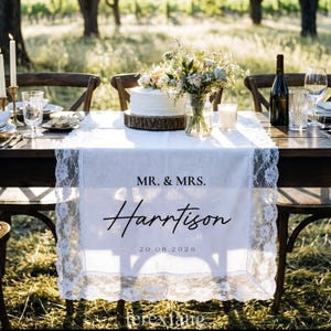 May include: A white linen table runner with lace trim, personalized with "MR. & MRS. Harrison" and the date 20.08.2026. The table is set for a wedding, with a cake, flowers, candles, and wine glasses.